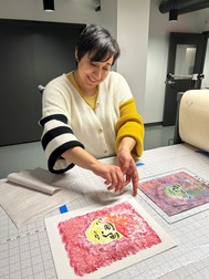 Artist smiles while preparing a colorful print on a worktable in a printmaking studio.