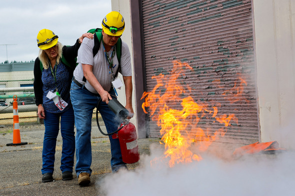 Two workers in hard hats use a fire extinguisher to put out flames during a workplace fire safety training.