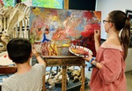 Child and instructor painting a colorful abstract canvas on an easel in an art studio with animal skeletons nearby.