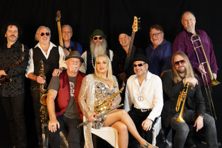 Large band posing with instruments on stage; a seated saxophonist in a gold dress is centered among musicians.