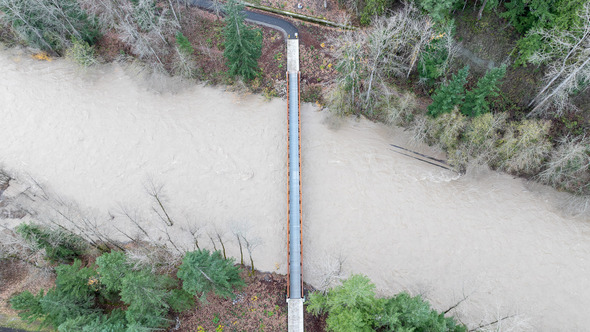 Aerial view of a pedestrian bridge over a swollen, muddy river with surrounding trees and high water levels.