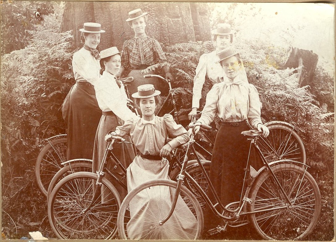 Vintage photo of five women in long skirts and hats posing with bicycles outdoors.