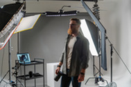 Photographer in a studio holding a camera, surrounded by lighting equipment and backdrops.