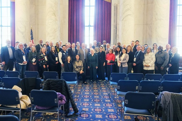 Large group of people posing in a formal chamber with tall columns, windows, and American flag.