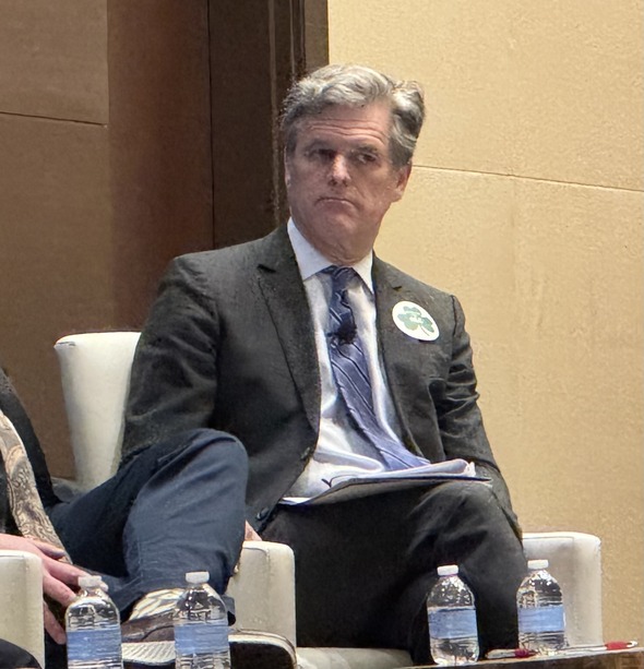 Man in suit seated on panel with microphone, holding notes, water bottles on table in front.