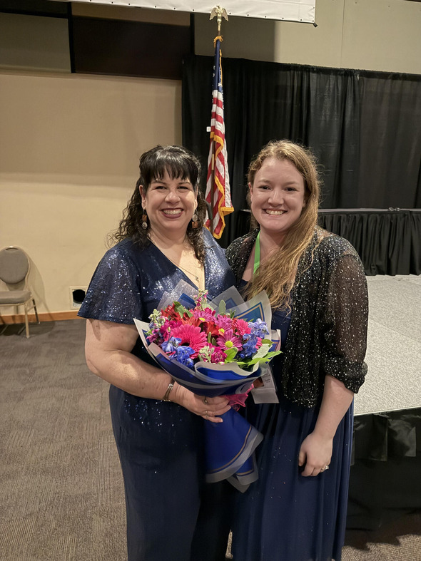 Two women smiling, holding a bouquet on stage with American flag in background.