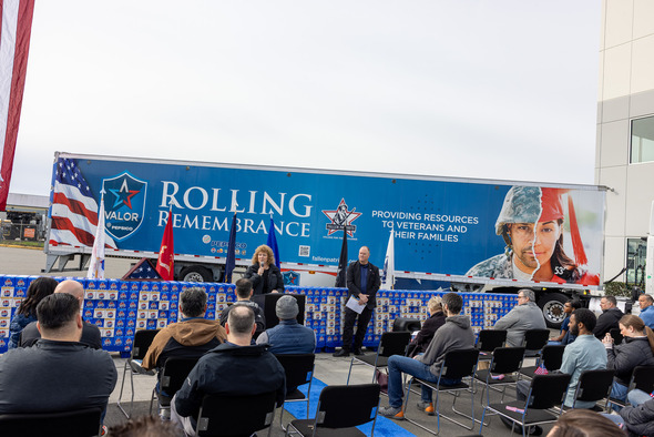 Speaker addresses seated crowd in front of Rolling Remembrance trailer highlighting support for veterans and families.