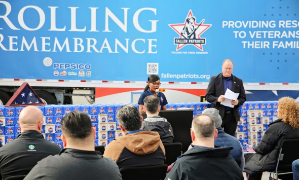 Speaker at podium during Rolling Remembrance event with audience seated in front of PepsiCo trailer honoring fallen veterans.