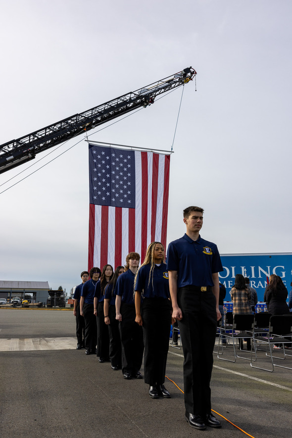 Students in uniform stand at attention beneath a large American flag suspended from a crane during ceremony.