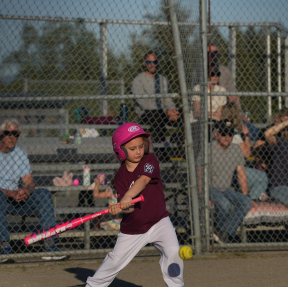 Young softball player in pink helmet swings bat as ball approaches, with spectators behind fence.
