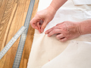 Hands pinning fabric pattern to cloth on a table with measuring rulers nearby.