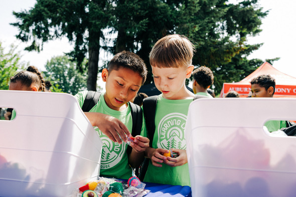 Two kids in green shirts examine small toys at an outdoor table during a community event.