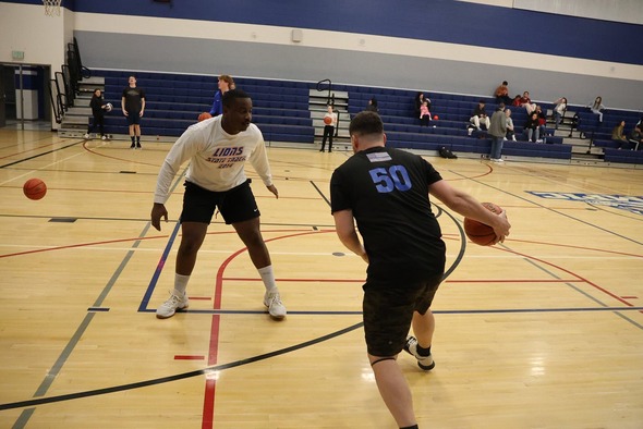 Two adults play basketball in a gym while others watch and practice in the background.