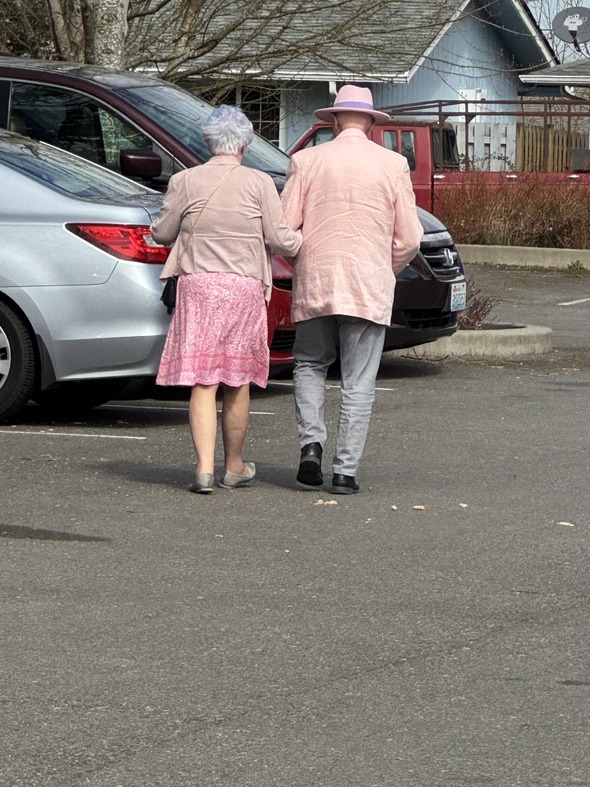 Elderly couple in pink outfits walk arm-in-arm through a parking lot, sharing a quiet, heartwarming moment.