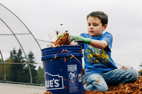 Child wearing gloves scoops mulch into a bucket outdoors, focused on helping with yard work.