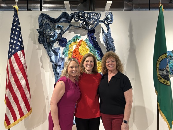 Three women stand smiling in front of artwork between U.S. and Washington state flags.