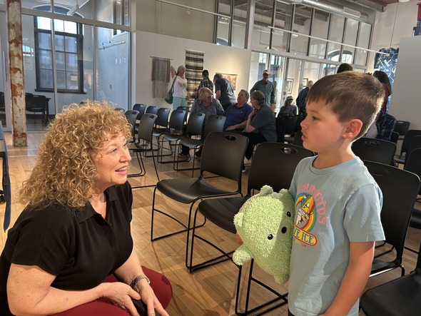 Woman kneels and smiles while talking to a young boy holding a green stuffed toy in a meeting room.