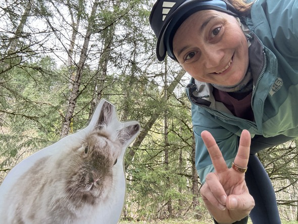 Person outdoors takes selfie with bunny cutout, flashing peace sign in wooded area.