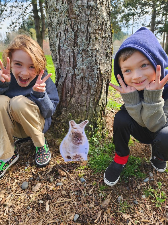 Two kids crouch by tree making peace signs next to a bunny cutout on the ground.