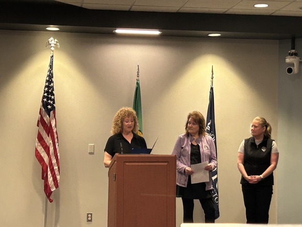 Mayor speaks at podium with two women nearby, flags behind them in a council or meeting room.