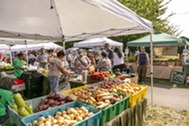 Busy outdoor farmers market with vendors and shoppers browsing fresh produce under white tents.