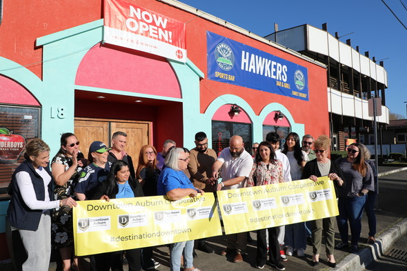 Ribbon cutting outside Hawkers Bar & Grill in Auburn, with crowd holding Downtown Auburn Cooperative banner under “Now Open” sign.