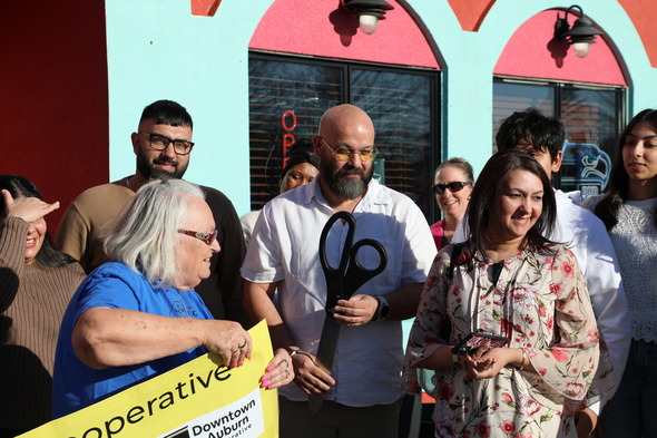 Close-up of ribbon cutting as man holds oversized scissors, smiling crowd gathered outside new Hawkers Bar & Grill.