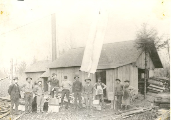 Vintage photo of workers standing outside a wooden mill building with large boards and stacked lumber.