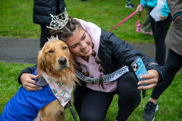 Teen wearing a sash and crown takes a selfie with a golden retriever dressed in a colorful outfit at an outdoor event.