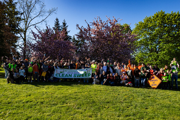 Large group of volunteers posing with a “Clean Sweep” banner in a park with blooming trees and sunshine.
