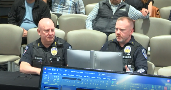 Two police officials present at a council meeting, seated behind a computer monitor, with audience members visible in the background.