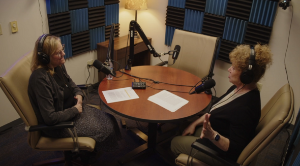 Two women sitting around a table with podcast mics and earphones on, they're recording a podcast in a cozy room 