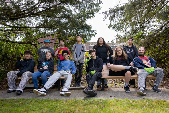 Group of teens sitting and standing around a newly built bench, smiling in a wooded area.