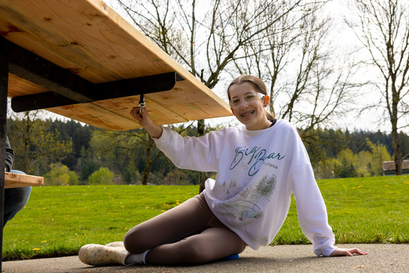Teen assembling a wooden bench outdoors, smiling while tightening hardware underneath.