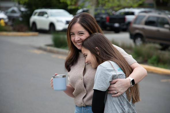 Woman hugging young girl outdoors, both smiling warmly in a parking lot setting.