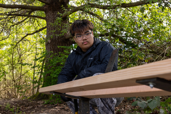 Teen working on bench assembly outdoors, focused while using tools near greenery.