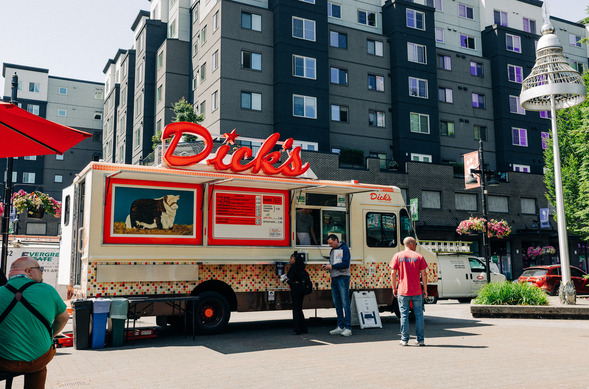 Dickās Drive-In food truck parked downtown with people ordering, surrounded by apartments and street planters.