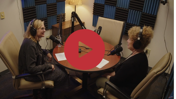 Two women wearing headphones record a podcast at a table with microphones inside a soundproof studio.