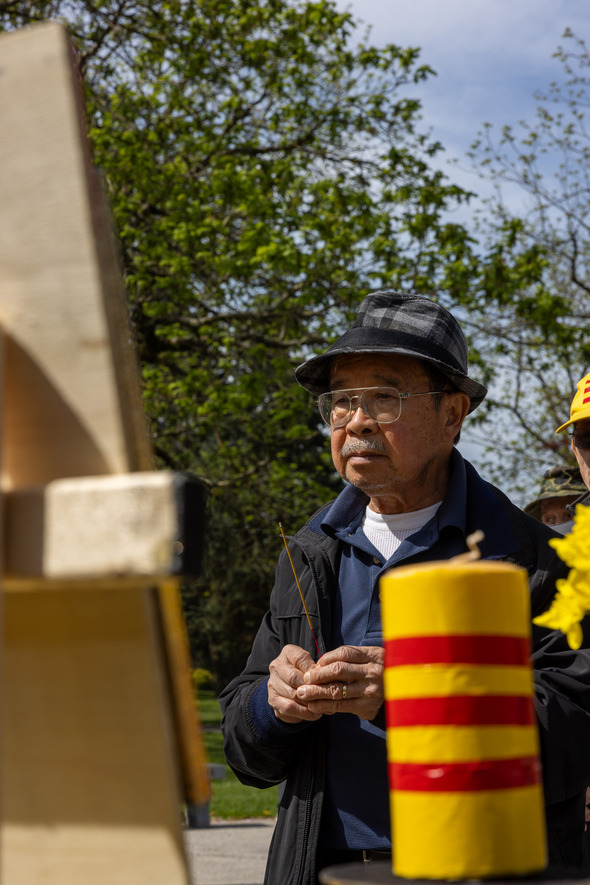 Older man holds incense during a memorial ceremony outdoors, with yellow flowers and a candle in the foreground.