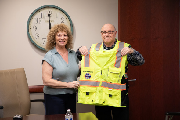 Mayor Backus and staff member smile while holding a high-visibility safety vest with city patch.