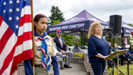 Woman speaks at a memorial event beside an American flag and scout, with attendees gathered under tents.
