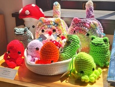 A group of crochet plush animals and objects in and out of a soft woven basket, all sitting on a wooden shelf