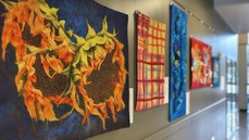 A quilted piece with sunflowers in main focus suspended on a wall in a row with other pieces