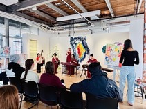 Band performing in front of a group of people seated with a person to the right standing in front of a painted canvas