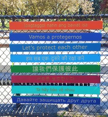 Eight multicolor wooden planks tied to a fence with the phrase "Let's protect each other" carved in different languages
