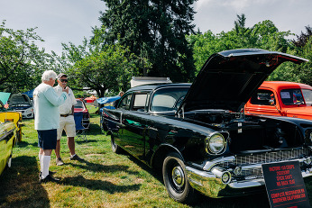 a factory 150 coupe 283/283HP sits on a field with the hood popped as onlookers discuss its features