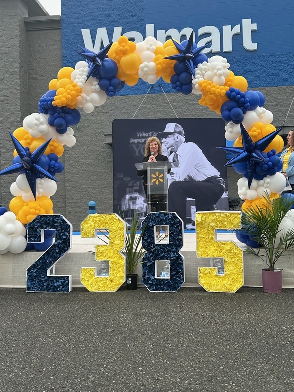 Speaker stands at a Walmart podium beneath a blue, yellow and white balloon arch.