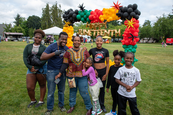 Family poses under a colorful Happy Juneteenth balloon arch at a park celebration.