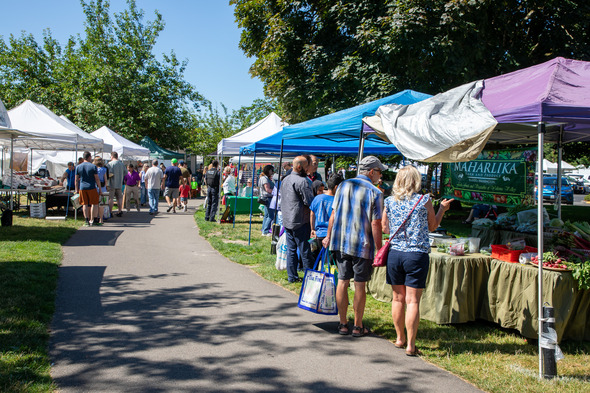 Shoppers walk through vendor tents at a sunny outdoor farmers market.