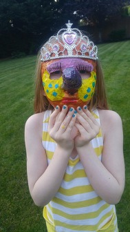 Child wearing a colorful handmade mask and tiara while standing outside on a lawn.